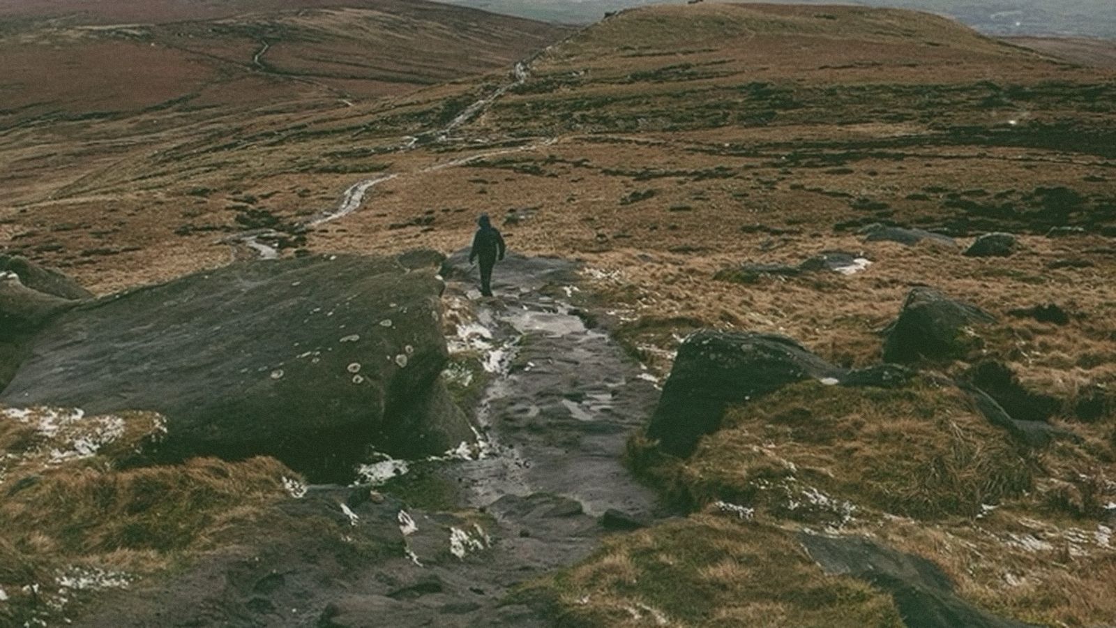 A lone walker on a wet gritstone path across brown moorland under a wide cloudy sky